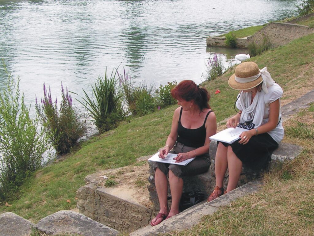 Dwie kobiety siedzące nad rzeką i rysujące z natury